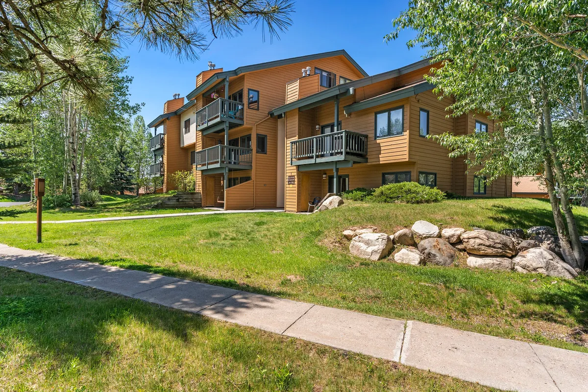 A serene exterior view with wooden balconies, natural stone landscaping, lush grass, and tall trees. The image captures residents walking and relaxing, further conveying the idea of a tranquil yet active community setting.