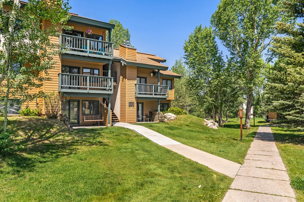 An exterior shot showing balconies with chairs, a pathway leading to the entrance, lush green grass, and surrounding trees. The image suggests a walkable, welcoming environment that blends nature and convenience.