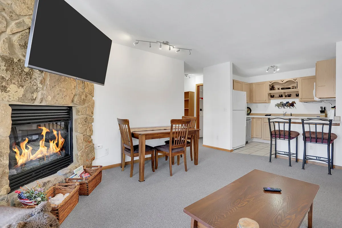 The image showcases a warm, inviting condo interior featuring a glowing gas fireplace as the centerpiece, a cozy dining table perfect for intimate meals or family gatherings, and a well-equipped kitchen area nearby. The scene evokes a sense of comfort and togetherness, ideal for relaxing after a day on the slopes or exploring downtown Steamboat Springs.