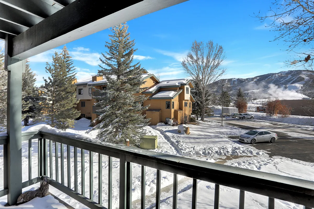 A scenic view of snow-covered ground, tall evergreen trees, wooden buildings, and a dramatic mountain backdrop. The image invites thoughts of winter sports and outdoor recreation, while the balcony hints at enjoying the landscape from the comfort of home.