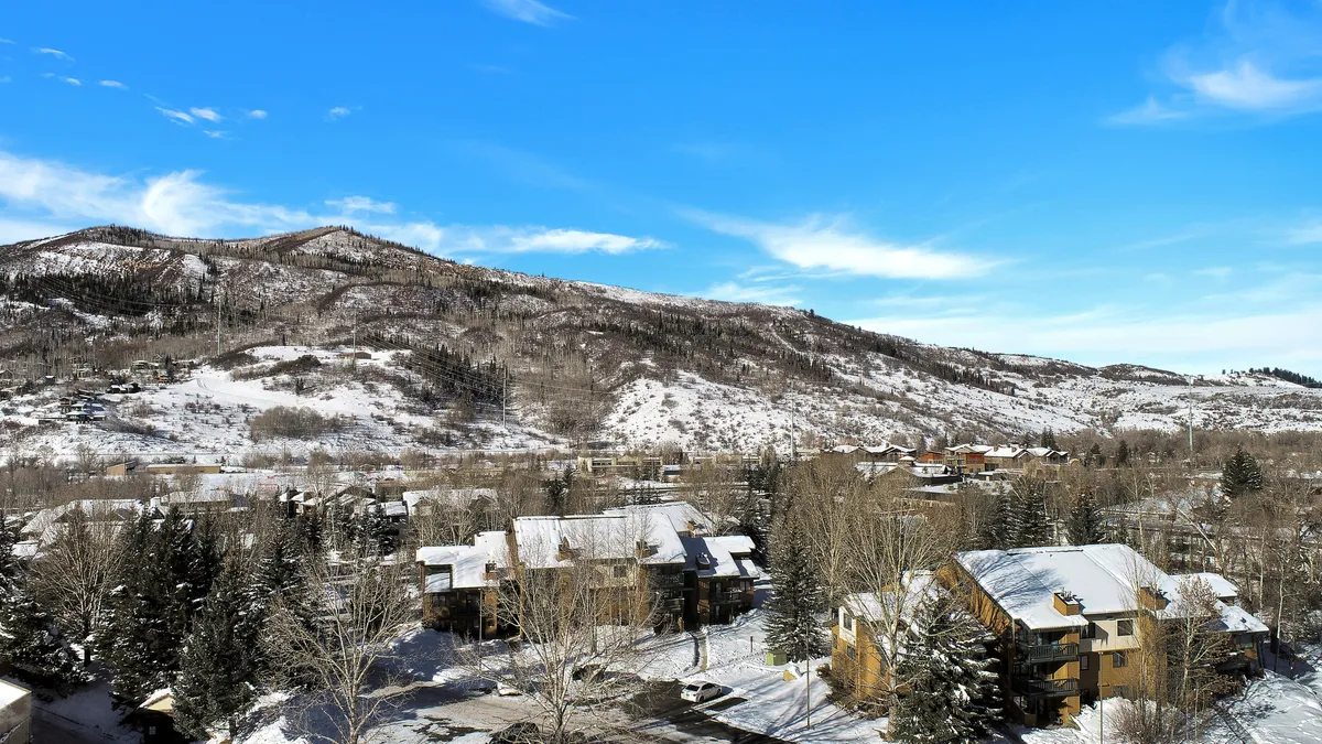 A tranquil winter scene featuring snow-covered mountains, residential buildings nestled among pine trees, and a wide expanse of snowy landscape. The image evokes the atmosphere of a ski town, with easy access to outdoor adventures visible just beyond the doorstep.