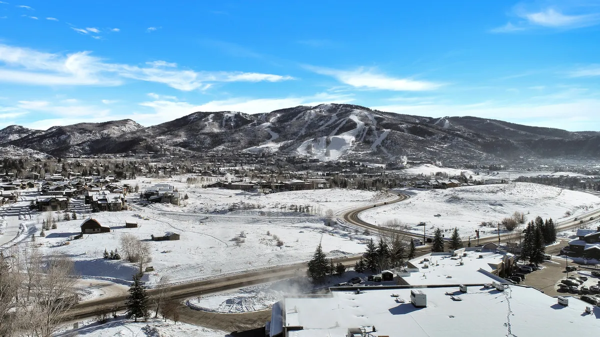 A sweeping view showing snow-covered mountains, a scenic valley, residential buildings, a winding road, and trees. The image captures the broader setting and highlights the blend of urban access and natural beauty.
