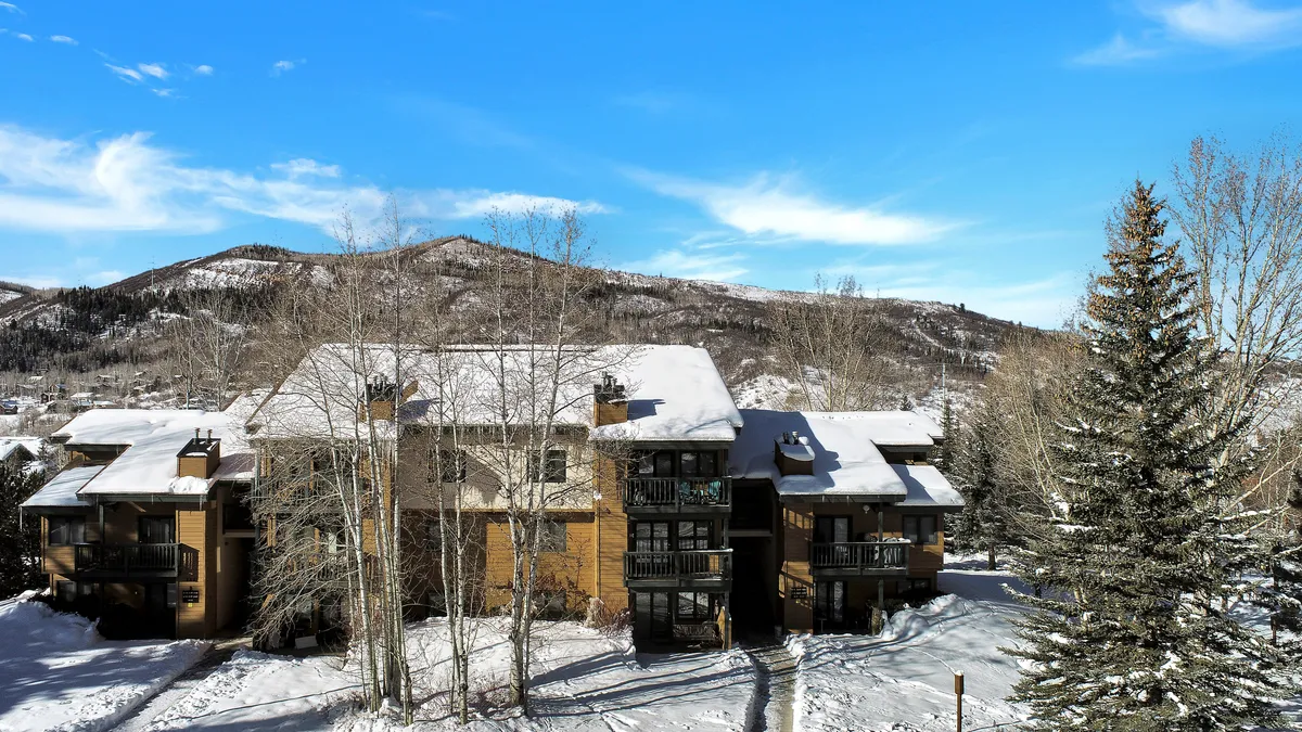 A snow-covered building nestled in a wintery landscape, flanked by tall evergreen trees and set against a backdrop of mountains. The scene captures the exterior of the condo, showcasing its inviting appearance and the natural beauty of the surrounding area.