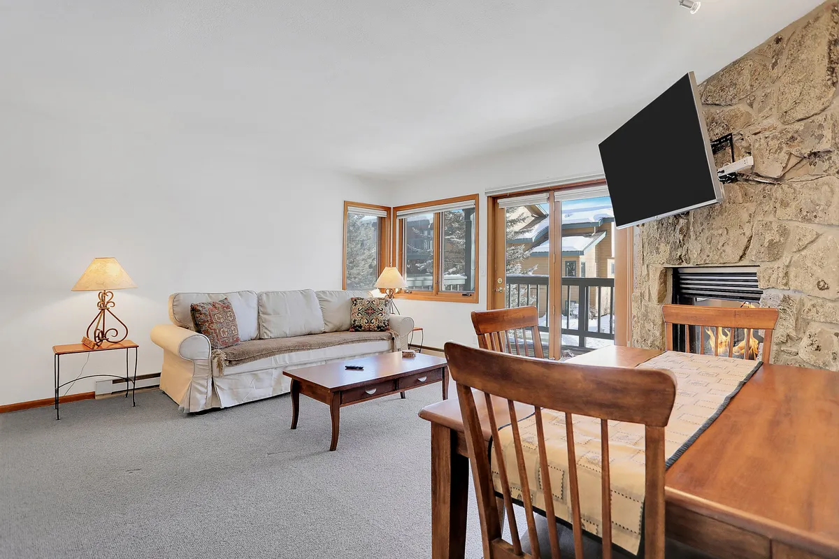 The photo presents a warmly lit living and dining area with a sofa facing the fireplace and a dining table nearby. Large windows let in natural light and reveal outdoor views, reinforcing the connection to the snowy mountain landscape.