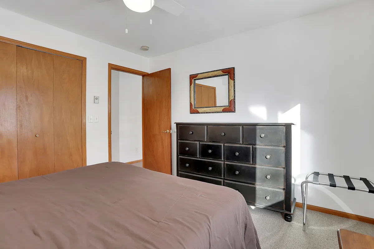 The image presents another angle of a bedroom, showcasing a neatly made bed, dresser, mirror, and doors. Natural light gently illuminates the space, emphasizing morning tranquility and preparation for the day ahead.